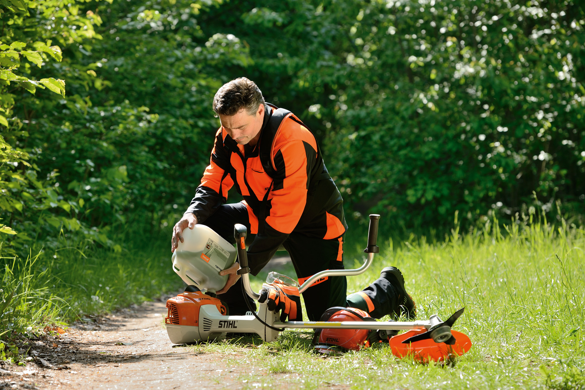 Um homem com um garrafão a abastecer o depósito de combustível de uma  roçadeira STIHL com arbustos atrás dele